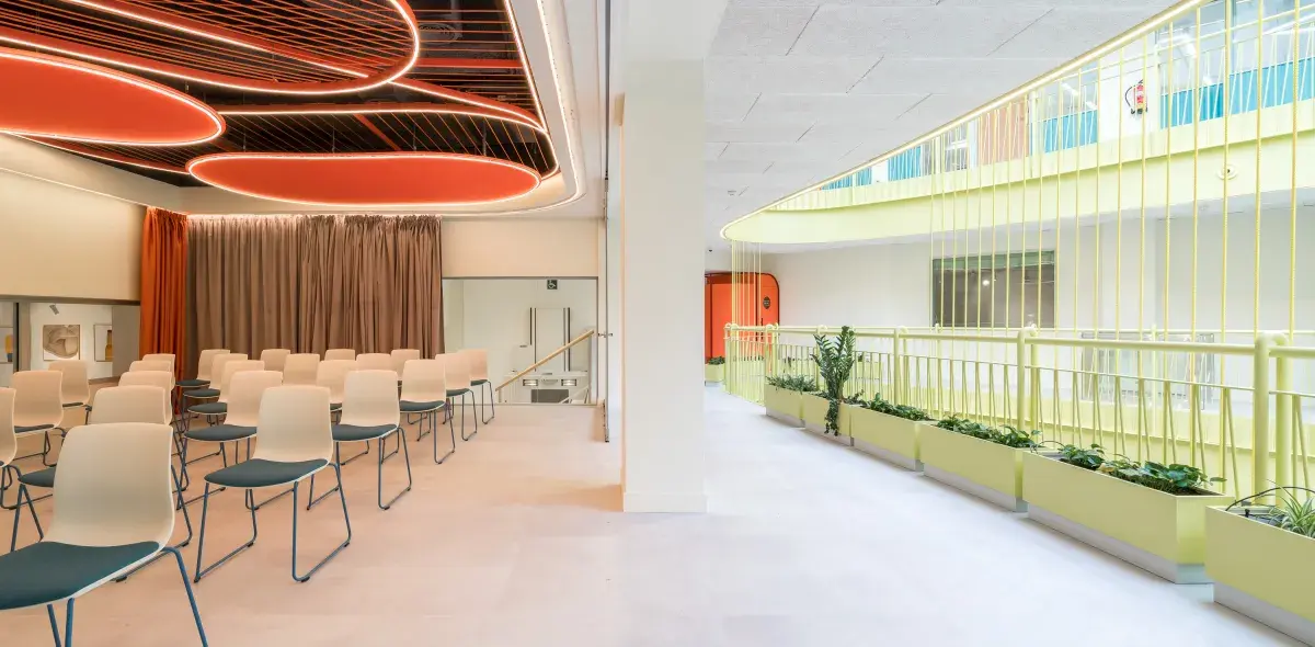 Sillas blancas y grises están dispuestas en una sala de conferencias, bajo un techo decorativo con luces naranjas, cerca de un pasillo interior con barandillas amarillas y plantas.