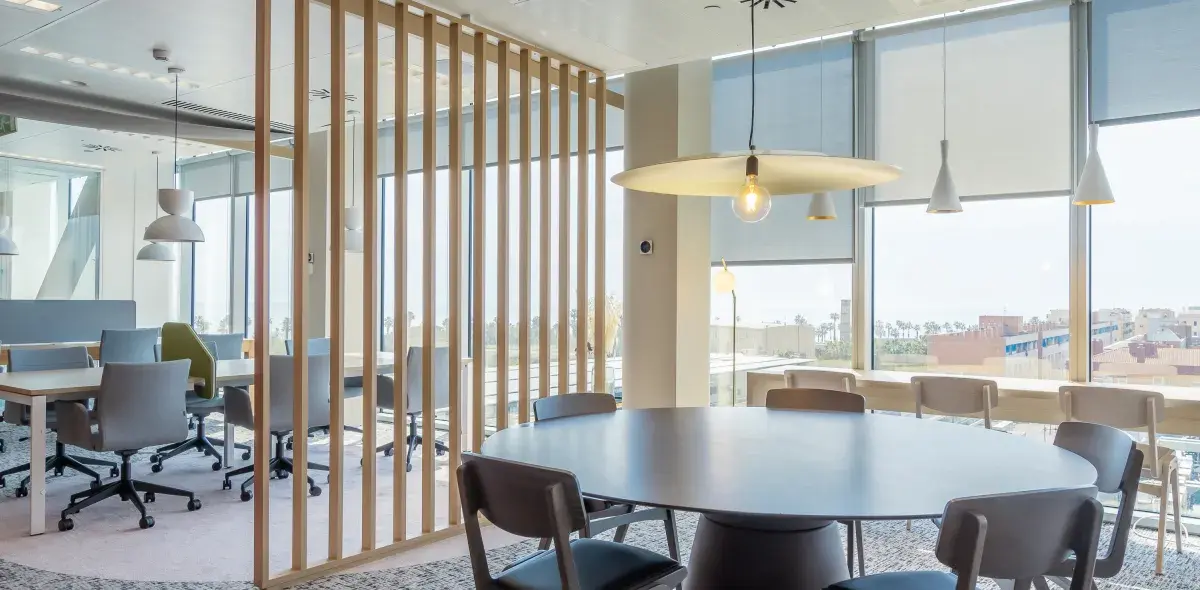 Una mesa redonda rodeada de sillas está en una sala de reuniones moderna con luminarias colgantes y ventanales grandes que ofrecen vista a un paisaje urbano.