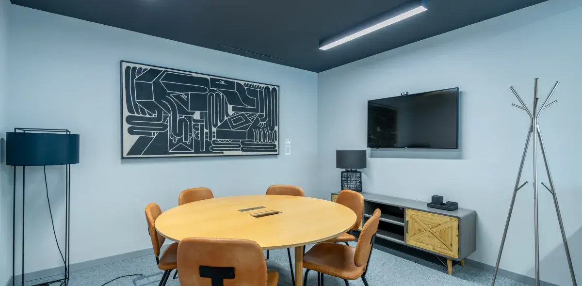Mesa redonda rodeada de sillas marrones en una sala moderna. En la pared, una obra de arte abstracto y un televisor. Lámpara de pie y perchero metálico al fondo.