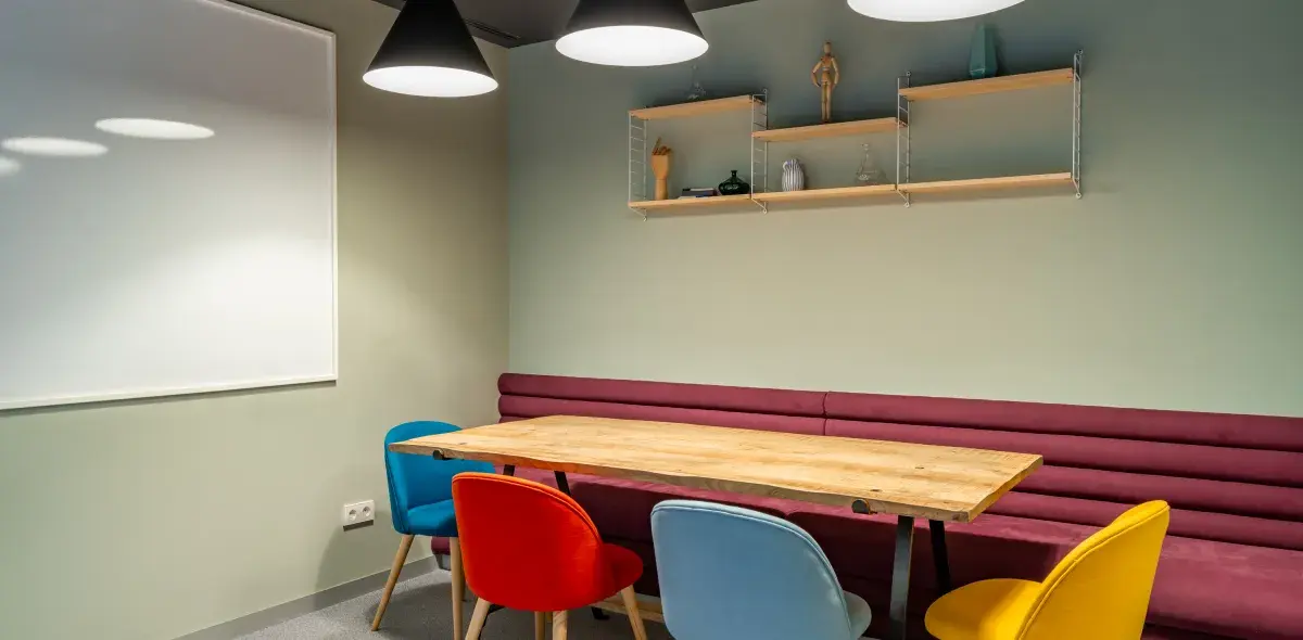 Mesa de madera rodeada de sillas coloridas, situada en una sala con luz colgante y pizarra blanca. Estanterías flotantes con decoraciones adornan la pared.
