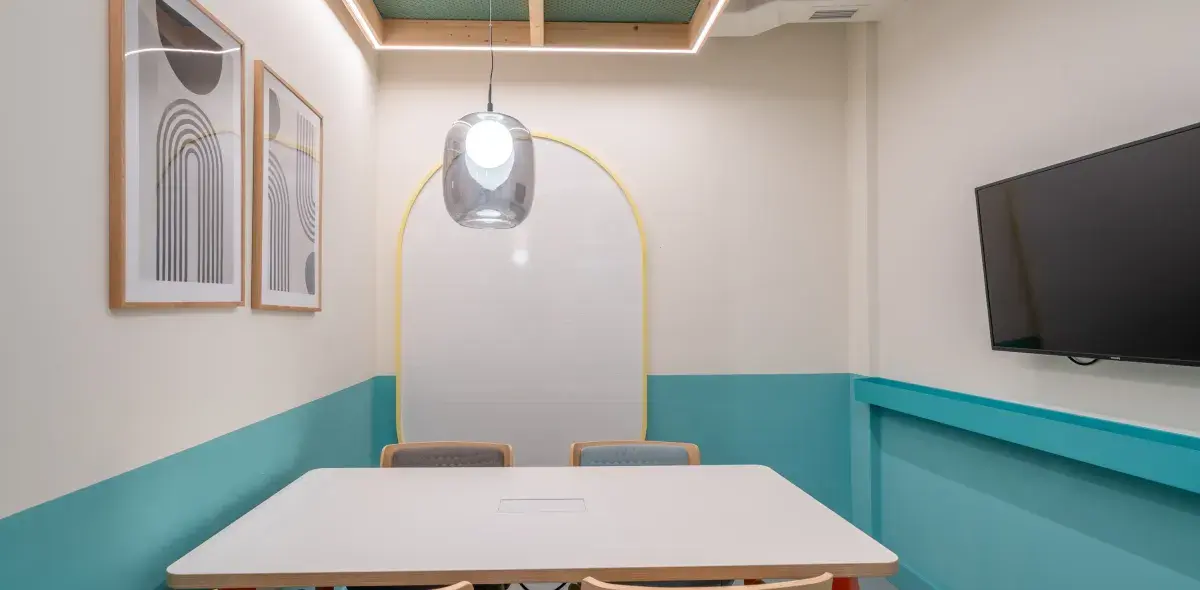 Una mesa blanca está en una sala de reuniones pequeña, iluminada por una lámpara moderna. Hay un televisor en la pared y tres cuadros abstractos.