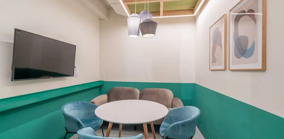 Una mesa circular blanca rodeada de sillas azul claro y un sofá gris en una sala pequeña con paredes turquesa. Una pantalla está colgada en la pared junto a dos cuadros abstractos.