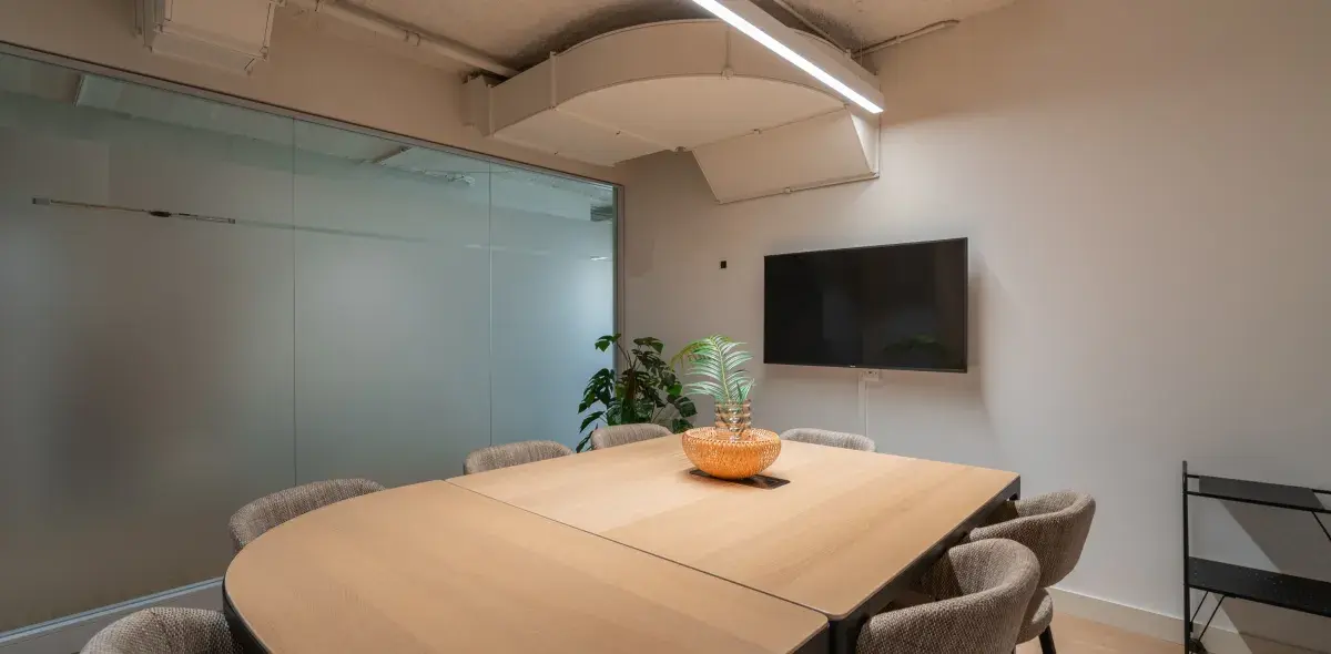 Mesa de reuniones con sillas alrededor; planta en jarrón como centro de mesa; pantallas en pared; sala con paredes de vidrio alrededor.