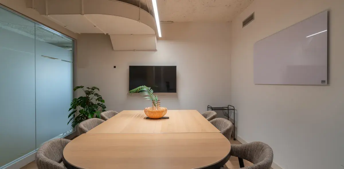 Mesa de madera rodeada de sillas, con planta decorativa encima, en una sala de reuniones moderna. Hay una pizarra blanca y una pantalla en la pared.