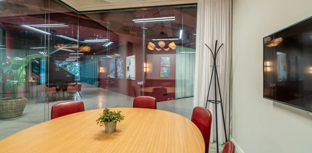 Mesa redonda de madera con sillas rojas rodeada de paredes de vidrio, vista a oficina moderna en un edificio con decoraciones y plantas.