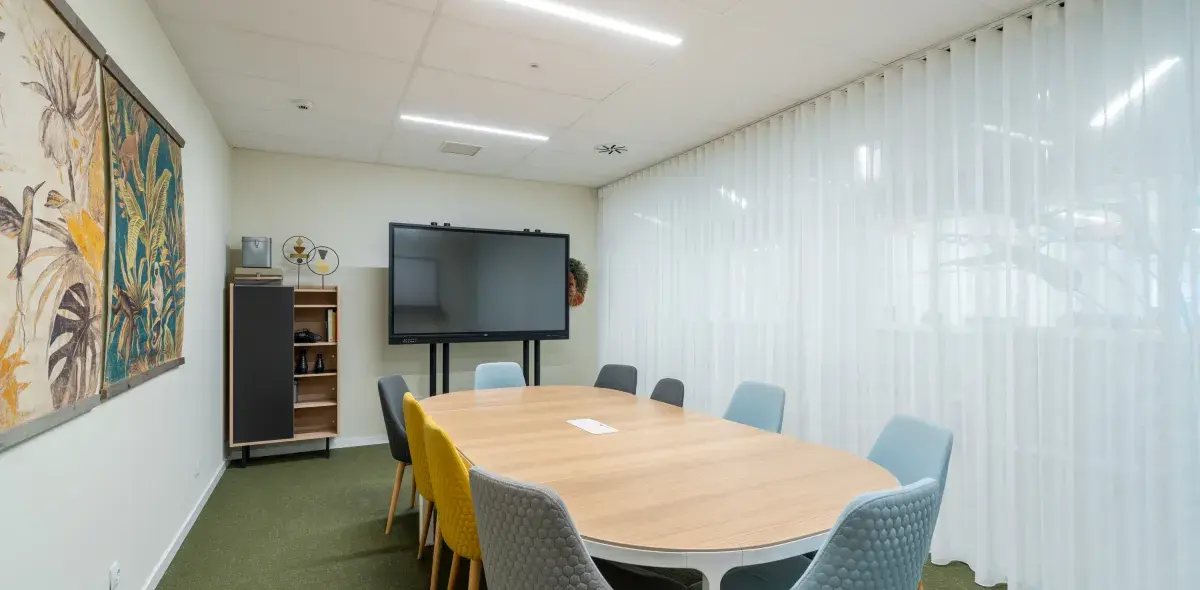 Una mesa ovalada rodeada de sillas de diferentes colores. En la pared, un monitor grande y arte decorativo, con cortinas blancas cubriendo ventanas.