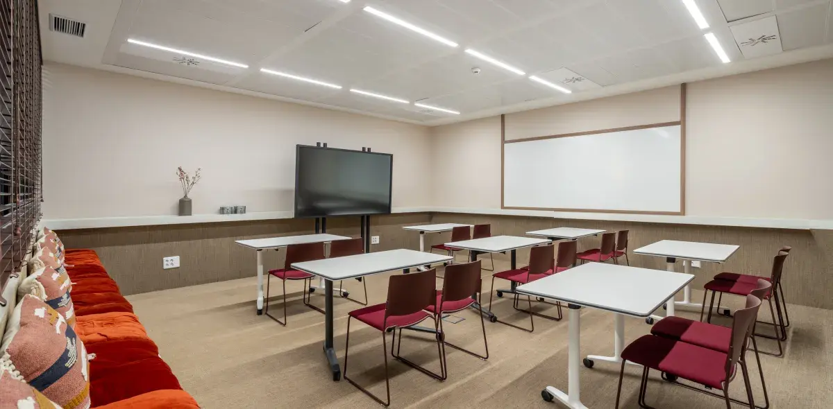 Una aula con mesas y sillas dispuestas en filas. Contexto: pantalla grande, pizarra blanca y cojines en un banco lateral. Ambiente moderno y bien iluminado.