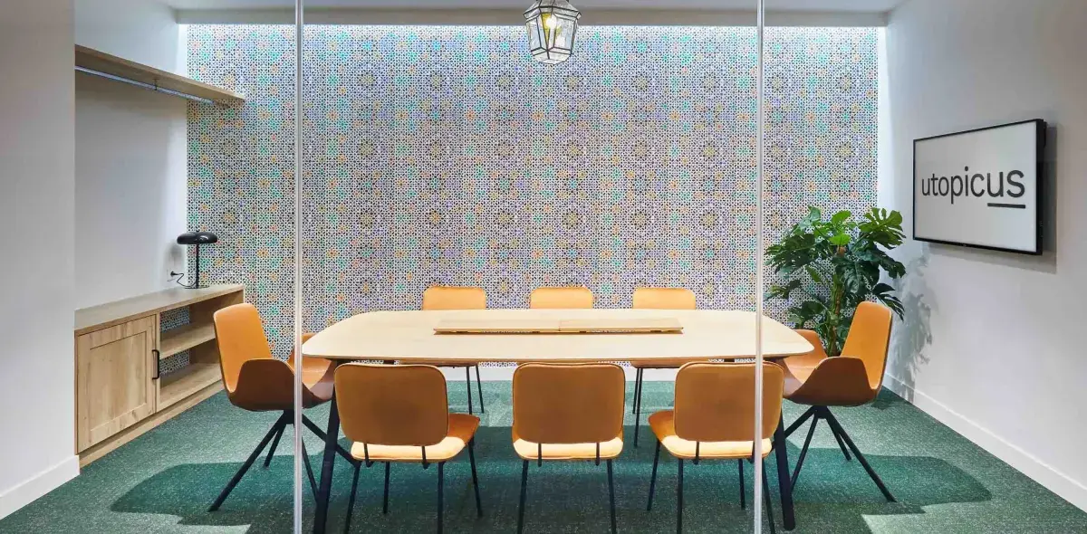 Una mesa ovalada rodeada de sillas anaranjadas está en una sala de reuniones moderna; en la pared, un cartel dice "utopicus".