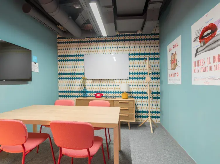 Una mesa de madera con sillas rojas en una sala de reuniones. Hay una pizarra blanca en la pared, carteles con textos "LA PIEL QUE HABITO" y "MUJERES AL BORDE DE UN ATAQUE DE NERVIOS".