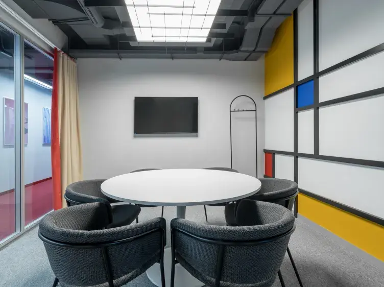 Una mesa redonda blanca rodeada de sillas negras se ubica en una sala de reuniones moderna con pared decorativa en colores primarios y una pantalla en la pared.