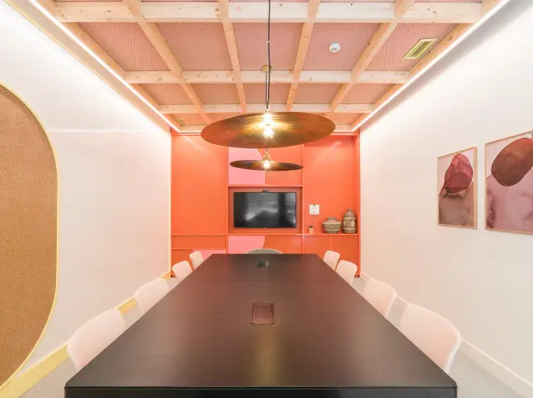 Una mesa rectangular negra con sillas blancas rodeada de paredes anaranjadas en una sala de reuniones con lámparas colgantes y un televisor en el fondo.