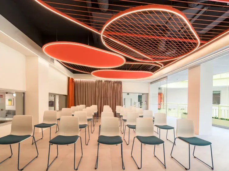 Filas de sillas vacías enfrentan una cortina marrón, en una sala moderna con techos decorativos iluminados en tonos naranja. Ventanas a la derecha permiten la entrada de luz natural.