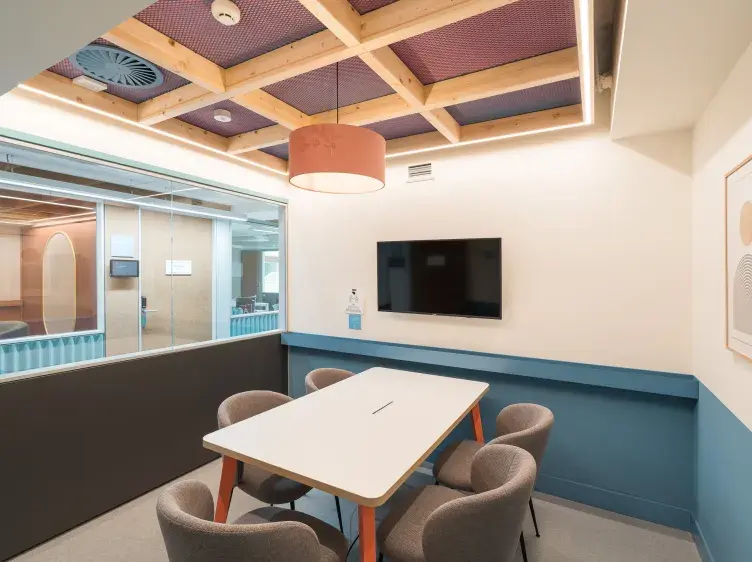 Una mesa blanca rodeada de sillas grises está en una sala de reuniones con pared de cristal, televisión montada y lámpara colgante en el techo.