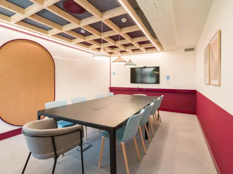 Una mesa rectangular negra con sillas azules alrededor, en una sala de reuniones moderna con paredes blancas y rojas. Hay una pantalla en la pared y lámparas colgantes.