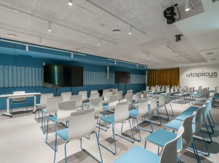 Sillas blancas alineadas en filas afrontan dos pantallas en una sala de conferencias moderna con paredes azules. El texto "utopicus" está visible en la pared derecha.