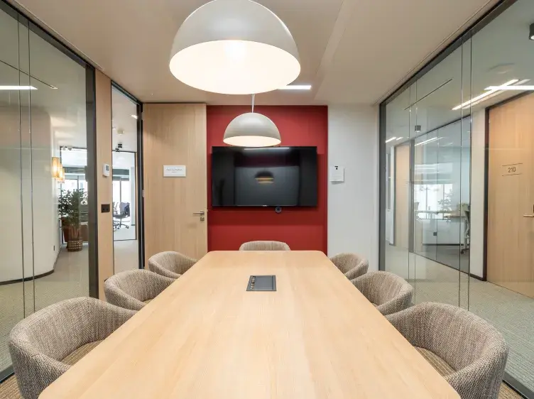 Mesa de madera en sala de reuniones, rodeada de sillas grises. Luces colgantes sobre la mesa, pantalla en pared roja. Puertas de vidrio a los lados.
