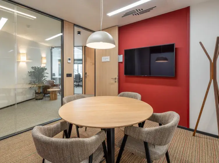 Mesa redonda con cuatro sillas rodeándola, situada bajo una lámpara colgante en una sala de reuniones moderna. Pared roja con pantalla de TV montada y perchero de madera.