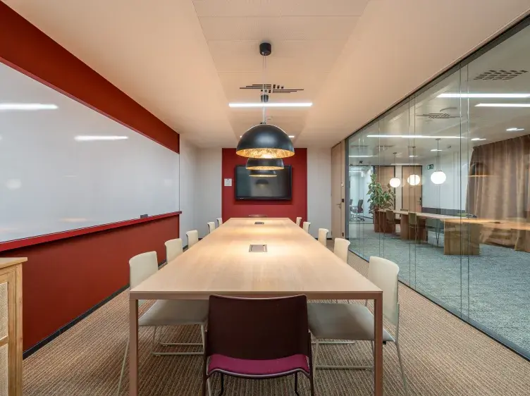 Una mesa rectangular rodeada de sillas está en el centro de una sala de reuniones con paredes de cristal, iluminación colgante y una pizarra blanca en la pared.