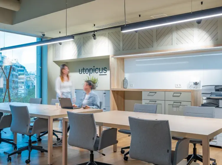 Personas trabajando en una oficina moderna con mesas largas y sillas; en la pared se lee "utopicus". Hay grandes ventanales que dejan ver edificios urbanos.