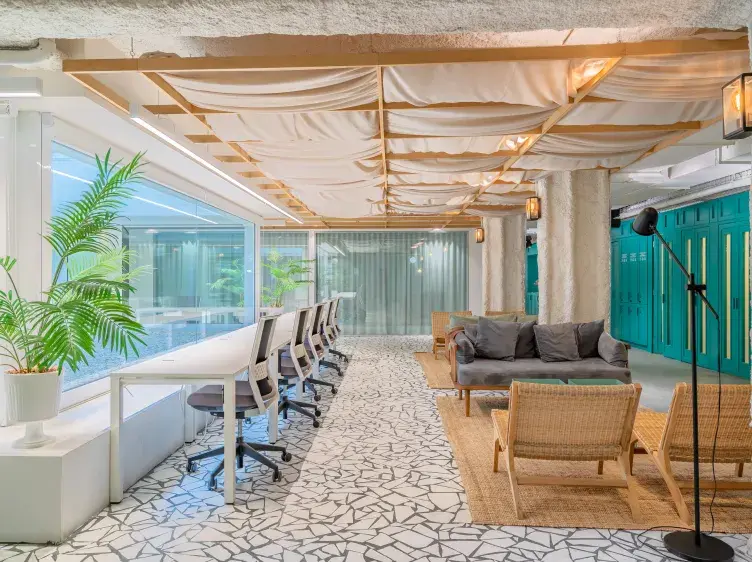 Espacio de oficina con escritorios alineados junto a una ventana, acompañados de sillas giratorias. A la derecha, hay sofás y sillas frente a casilleros turquesas. Decoración moderna y luminosa.