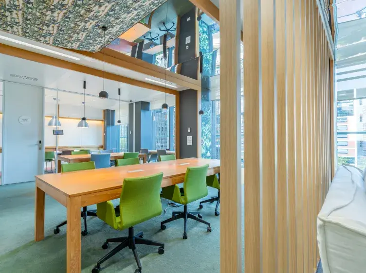 Mesa de trabajo con sillas verdes giratorias, en una oficina moderna con paredes de vidrio y lámparas colgantes. Se observan edificios urbanos a través de las ventanas.