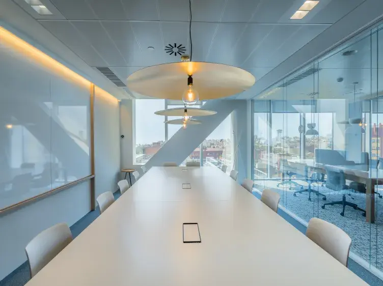 Mesa larga blanca con sillas, lámparas colgantes, situada en una sala de reuniones con ventana panorámica y paredes de vidrio. Al fondo, se observan edificios y cielo despejado.