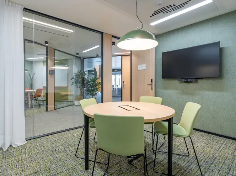 Una mesa redonda con sillas verdes está en el centro de una sala de reuniones moderna con paredes de vidrio y una pantalla de televisión montada en la pared.
