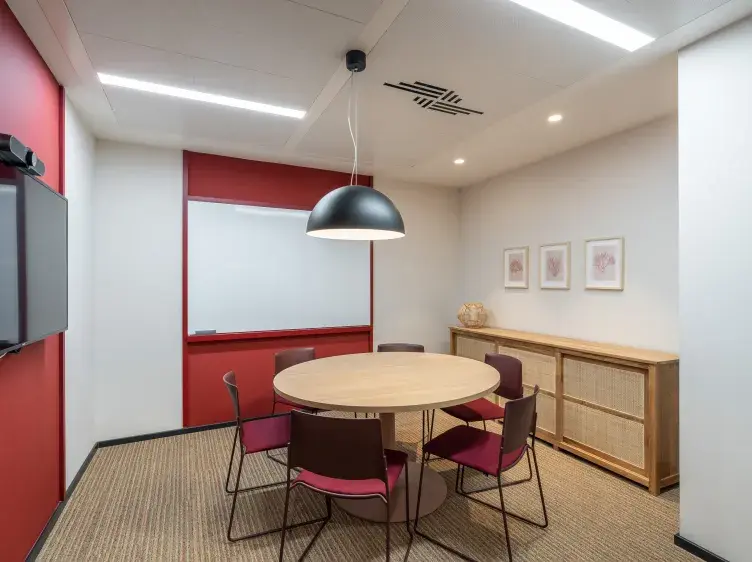 Mesa redonda con seis sillas alrededor, bajo una lámpara colgante negra; contexto de oficina con pizarra, televisor, y muebles de madera. Tres cuadros decoran la pared.