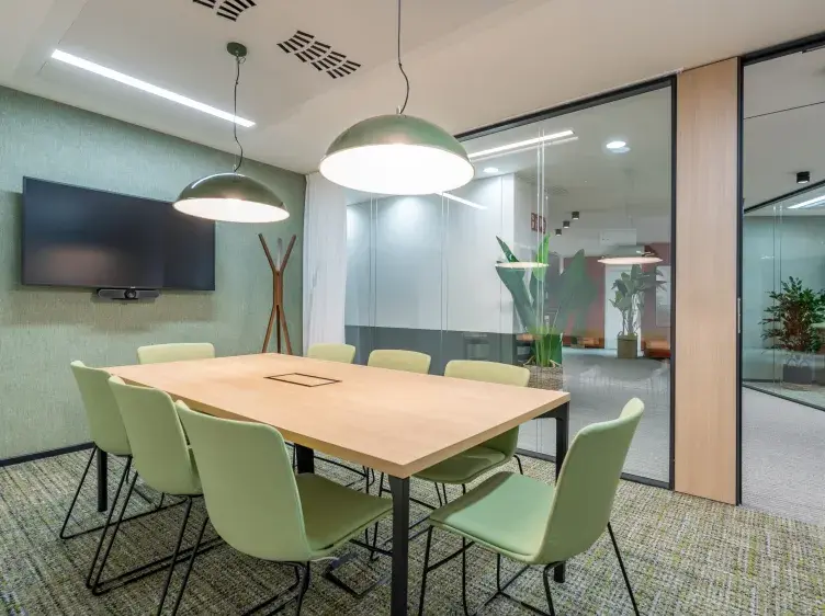 Mesa rectangular rodeada de sillas verdes, iluminada por lámparas colgantes, en una sala de reuniones con televisor y paredes de vidrio.