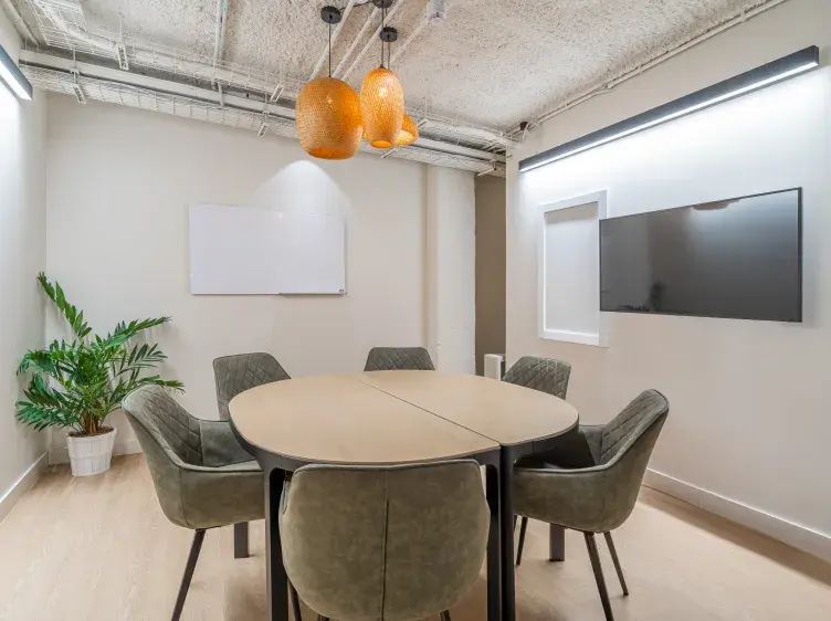 Una mesa redonda rodeada de sillas está en una sala de reuniones, iluminada por lámparas colgantes. Un televisor y una pizarra adornan la pared blanca; planta en la esquina.