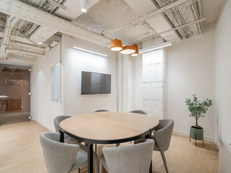 Una mesa redonda rodeada de sillas, ubicada en una sala de reuniones moderna. En la pared, hay una pantalla montada y una planta en una esquina. Al fondo, una puerta de vidrio con texto que dice "RECADIA ROOM".