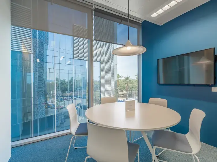 Una mesa redonda blanca rodeada de sillas está en una sala de reuniones moderna, con ventanas grandes y una pantalla en la pared azul.