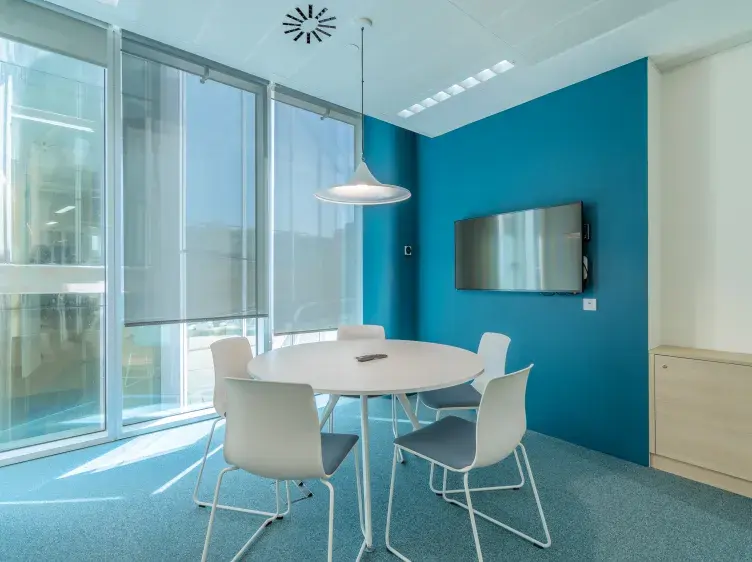 Mesa redonda blanca rodeada de cinco sillas en una sala con paredes azules y ventana amplia. Hay una lámpara colgante sobre la mesa y una pantalla en la pared.