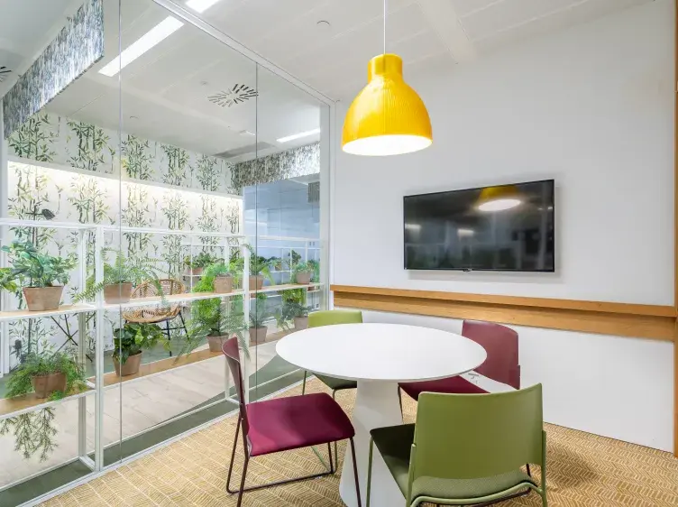 Una mesa redonda blanca con sillas de colores está en una sala moderna, iluminada por una lámpara amarilla. Al fondo, una pared de vidrio muestra plantas en macetas.