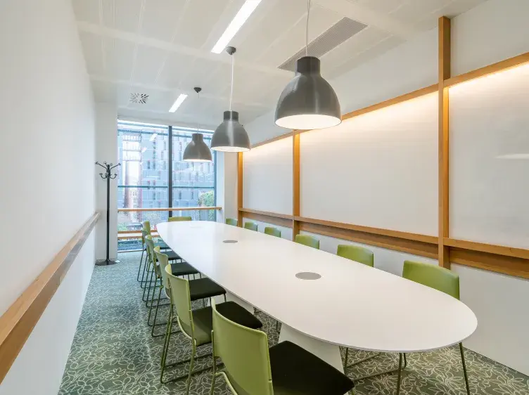 Mesa ovalada blanca con sillas verdes, rodeada de lámparas colgantes en una sala de reuniones moderna, con ventanas grandes y paredes blancas.