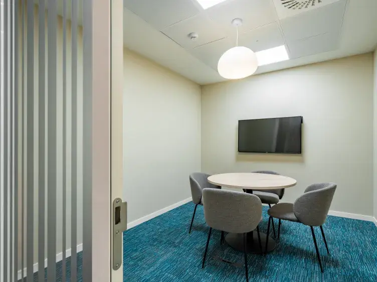 Una mesa redonda con cuatro sillas grises está en una pequeña sala de reuniones. Hay una pantalla en la pared y alfombra azul brillante en el suelo.