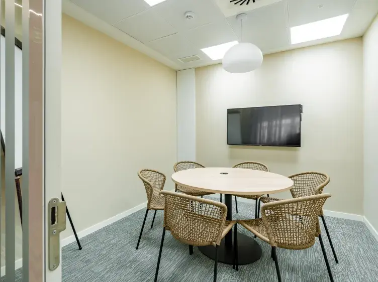 Mesa redonda rodeada de sillas tejidas, en una sala de reuniones con televisión montada en la pared y luz colgante.