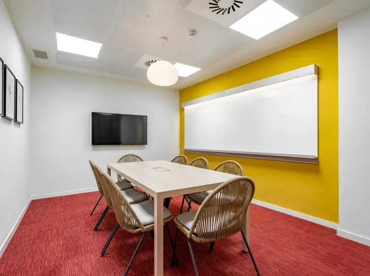 Mesa con sillas rodeándola, situada en una sala de reuniones; fondo amarillo con pizarra y televisión, alfombra roja.