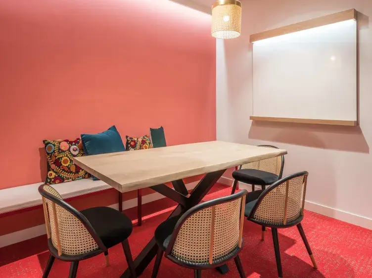 Mesa rectangular de madera con sillas alrededor, situada en una sala con pared rosa, cojines y pizarra blanca. Ambiente iluminado por lámpara de techo.