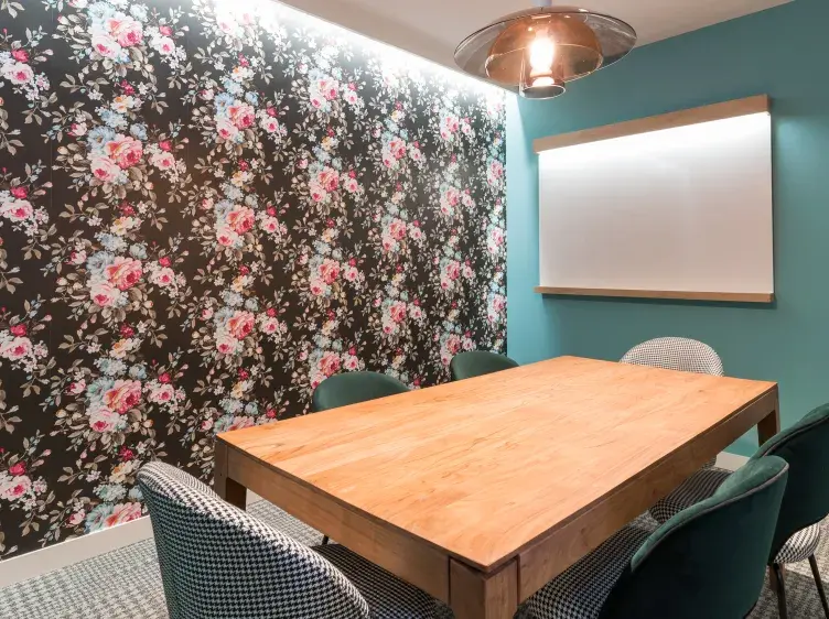 Mesa de madera rodeada de sillas en una sala de reuniones con pared floral y pizarra blanca iluminada.