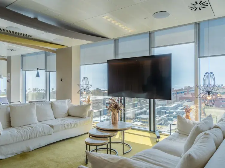 Sofás blancos se enfrentan, con una mesa circular entre ellos, en una sala con ventanas grandes y vistas urbanas. Una pantalla grande está montada en la pared. Iluminación moderna y decorativa.
