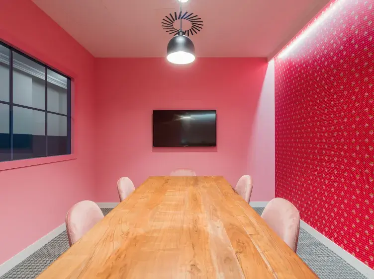 Mesa de madera rodeada de sillas rosadas, bajo una lámpara negra; ambiente de paredes rosas y una televisión montada. Ventana a la izquierda y pared decorativa roja a la derecha.