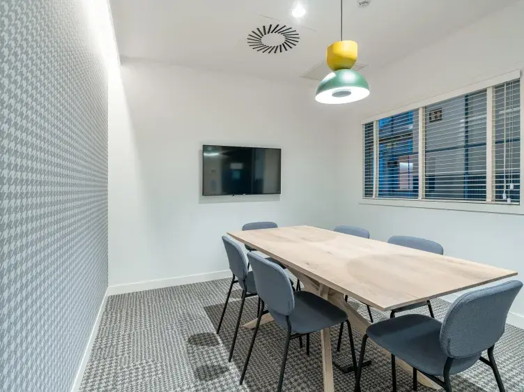 Una mesa con sillas rodea un televisor montado en la pared, bajo una lámpara colgante, en una sala de reuniones.