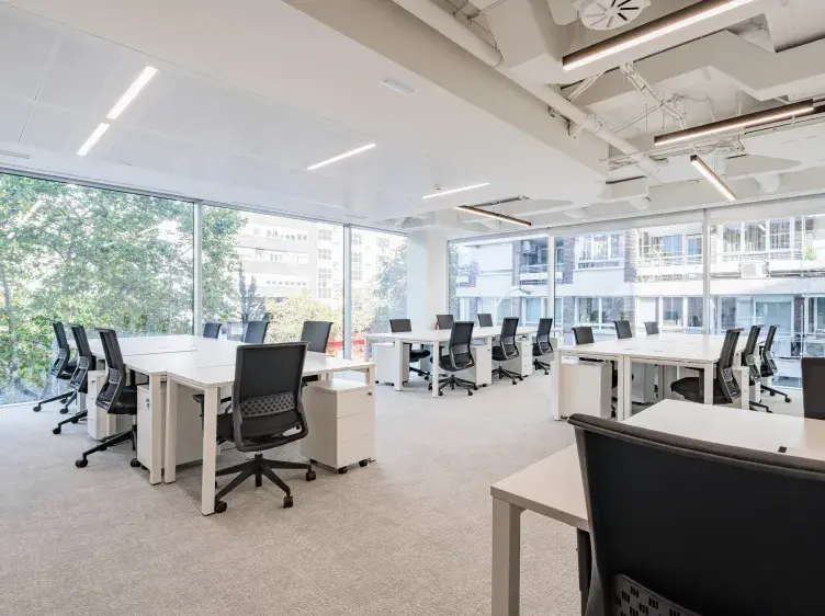Oficina con escritorios blancos y sillas negras distribuidas en un espacio amplio y moderno, junto a grandes ventanales que ofrecen vistas al exterior urbano.
