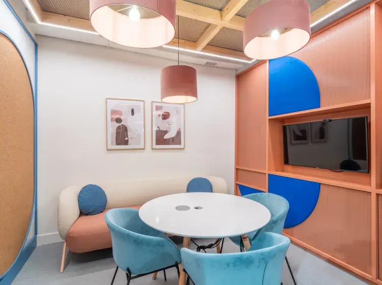 Una mesa blanca redonda rodeada de sillas azules en una sala moderna con sofá, arte en la pared y lámparas colgantes. Al fondo, estantes geométricos color naranja y azul.