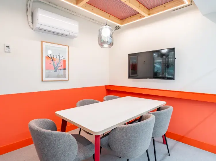 Mesa alargada rodeada de sillas grises, situada en una sala con paredes blancas y naranja. Hay un aire acondicionado, un televisor en la pared y un cuadro decorativo.