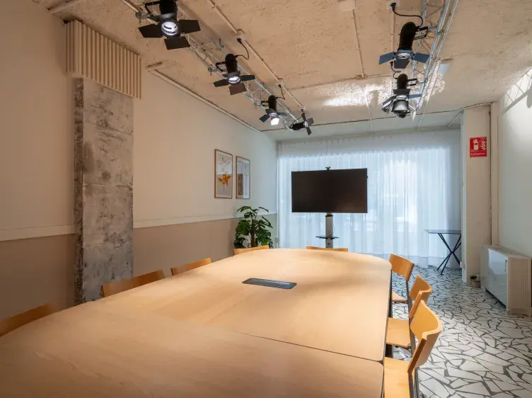 Una mesa de reuniones está rodeada de sillas, con un televisor frente a ella; en un salón iluminado, decorado con plantas, cuadros y luces en el techo.