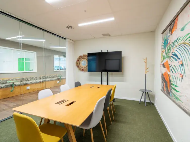 Mesa de madera con sillas alrededor en una sala de reuniones. Paredes con pintura, televisión y decoración.