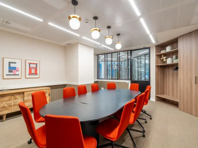 Una mesa ovalada negra rodeada de sillas naranjas en una sala de reuniones, con luces colgantes y estantes de madera en la esquina. En la pared, dos cuadros geométricos.