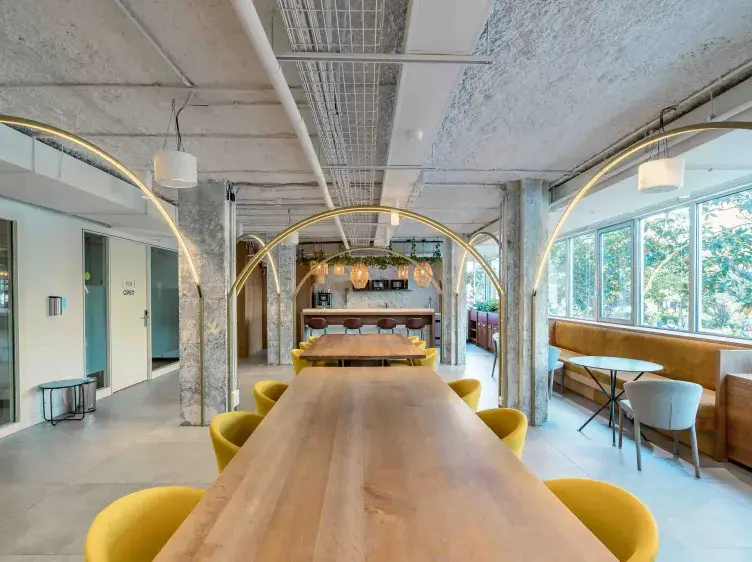 Una mesa de madera alargada rodeada de sillas amarillas, en un espacio moderno con arcos dorados y grandes ventanales. Al fondo, una barra con plantas colgantes y lámparas decorativas.